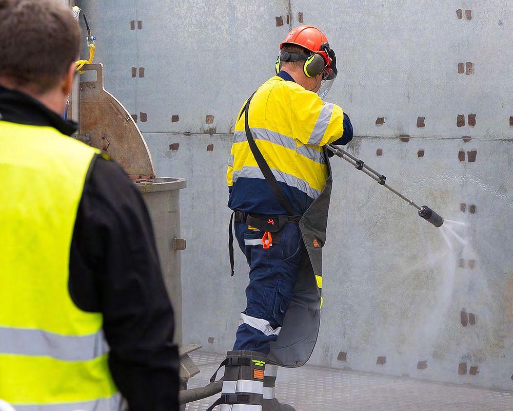 Operator in hi-vis and protective gear using a high-pressure water lance on a steel wall