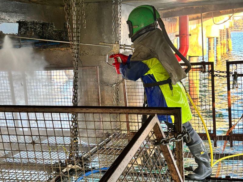 Aqua Prep operator in a green blasting helmet and protective gear water blasting a concrete structure over the water